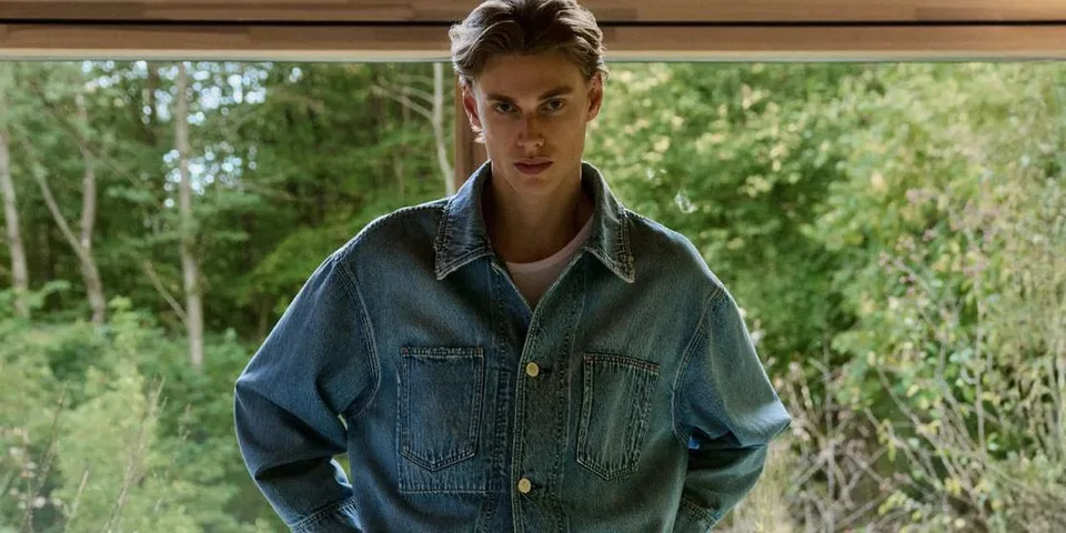 Homme en veste en jean devant une fenêtre, regardant la caméra.