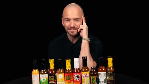Homme avec plusieurs bouteilles de sauce piquante alignées sur une table.