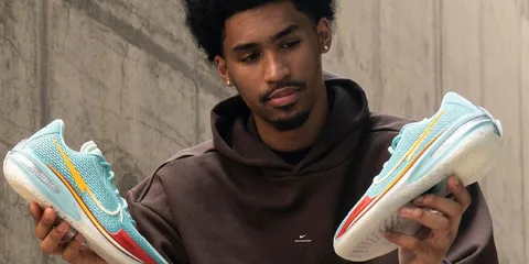 Homme en pull marron tenant des baskets colorées, fond urbain gris.