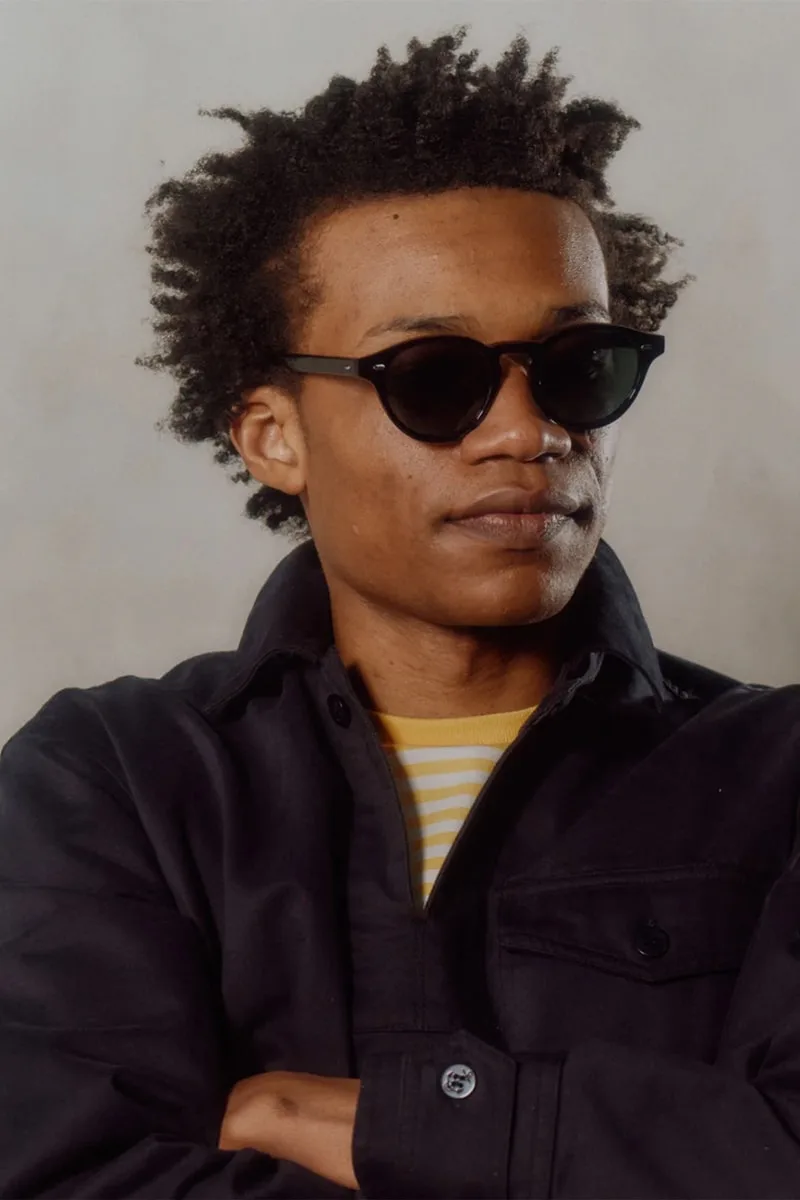 Un homme avec des lunettes de soleil et une veste noire regarde ailleurs.