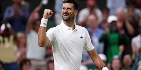 Homme en tenue de tennis blanc levant le poing, foule floue en arrière-plan.