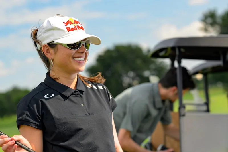 Femme en casquette blanche souriante avec lunettes de soleil sur un terrain de golf.