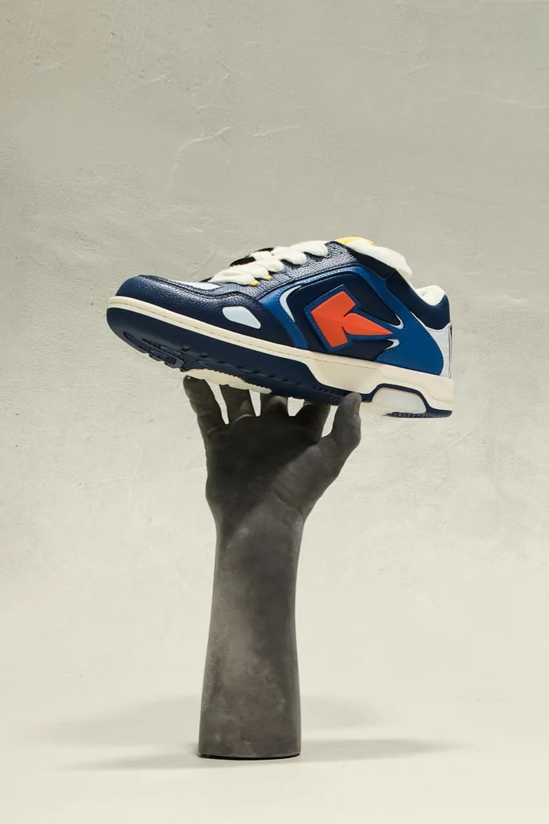 Main tenant une basket bleue avec logo rouge, fond blanc gris.