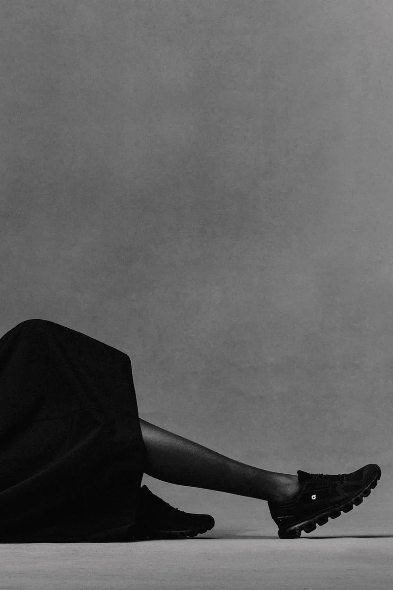 Personne allongée, jupe noire et baskets, fond gris.