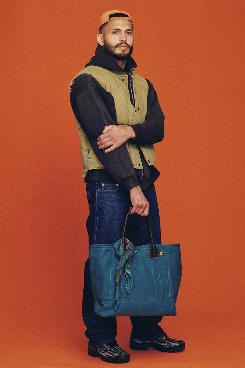 Homme posant avec un sac bleu sur fond orange, veste et casquette.