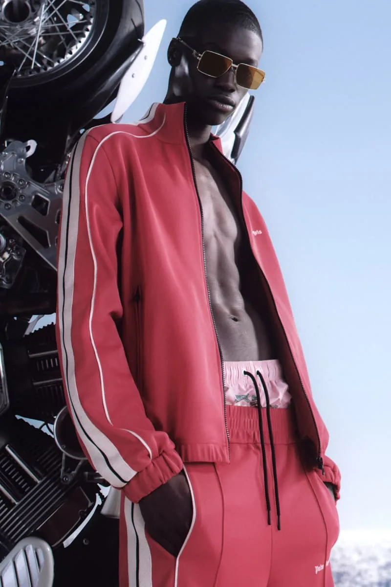 Homme en tenue sport rouge devant une moto et un ciel bleu.