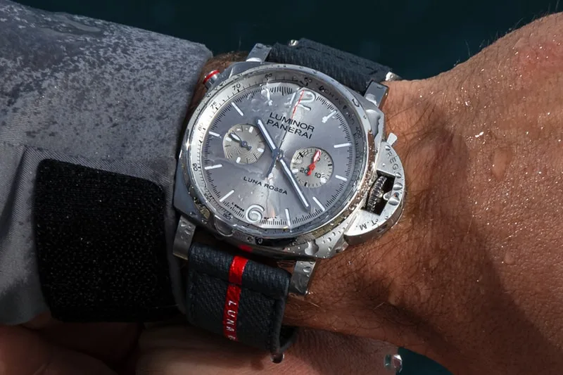 Montre argentée avec bracelet noir et détails rouges, sous la pluie.