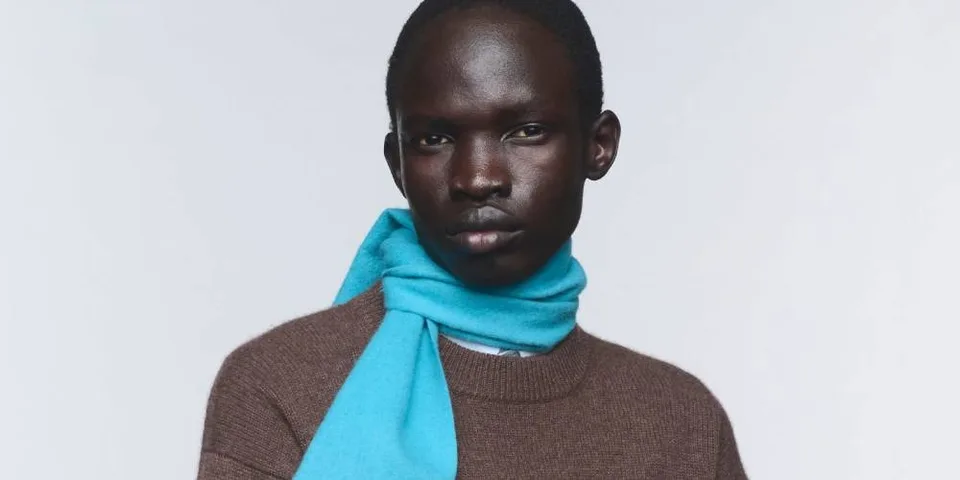 Personne avec écharpe bleue et pull marron, fond blanc.