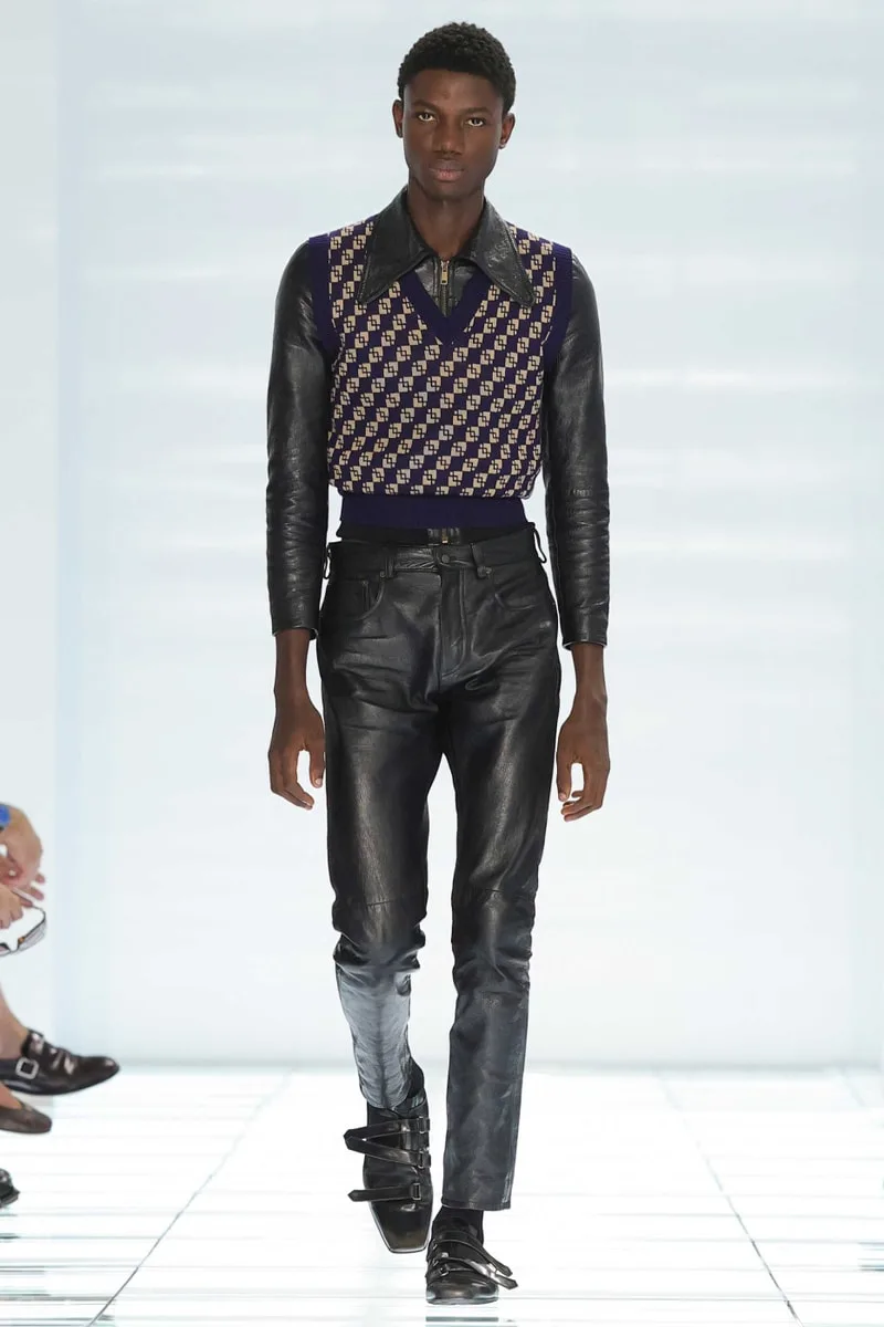 Homme défilant en tenue chic et moderne avec pull violet à motifs et pantalon en cuir.