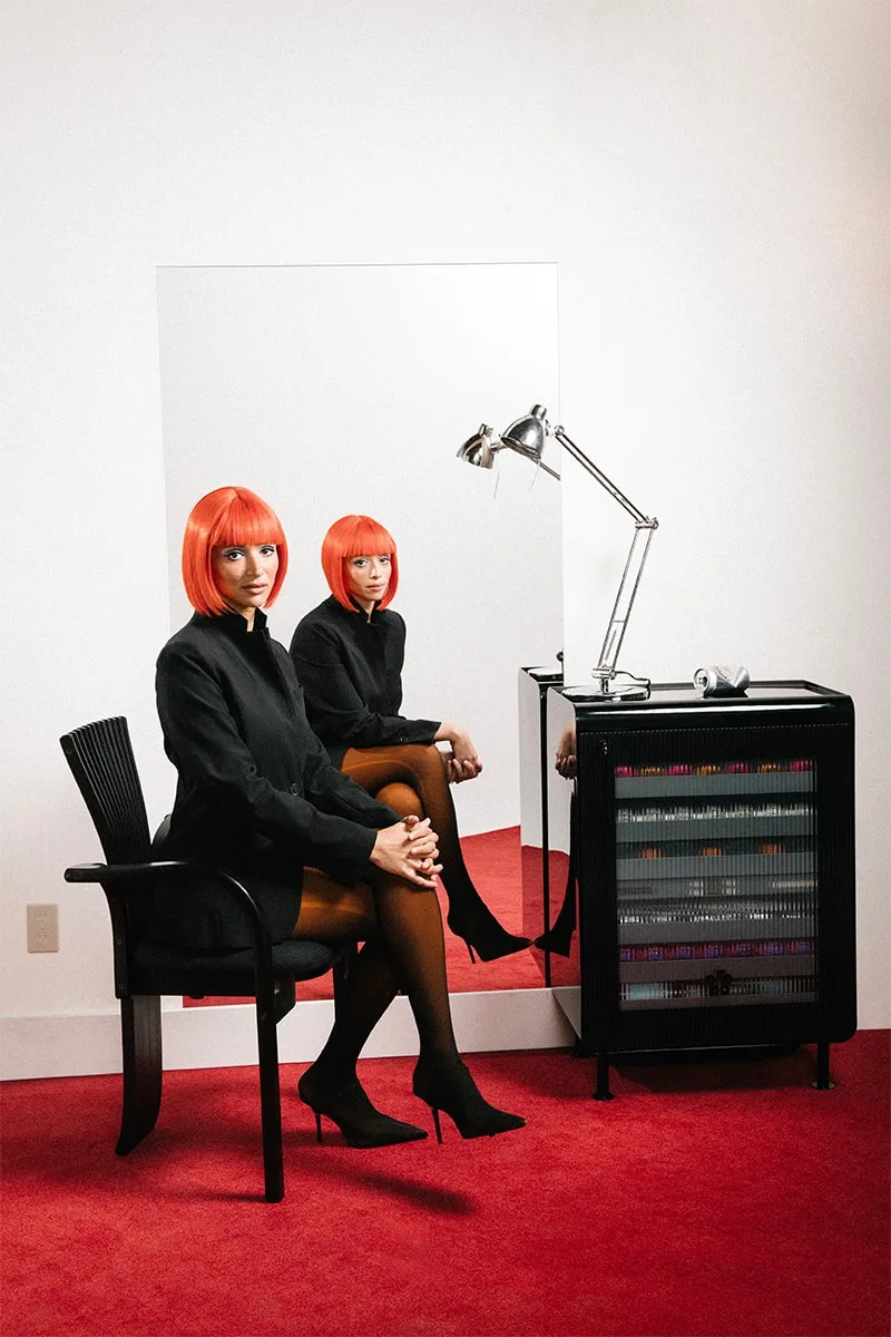 Femme en perruque rouge posant devant un miroir, intérieur moderne.