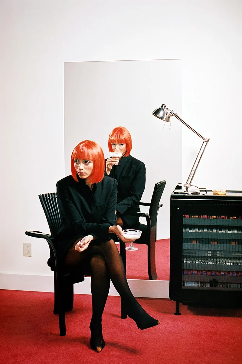 Femme rousse en noir devant un miroir, pièce rouge et blanche.