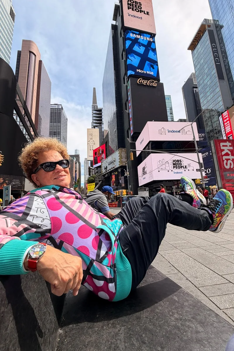Homme assis à Times Square, vêtements colorés et lunettes de soleil.