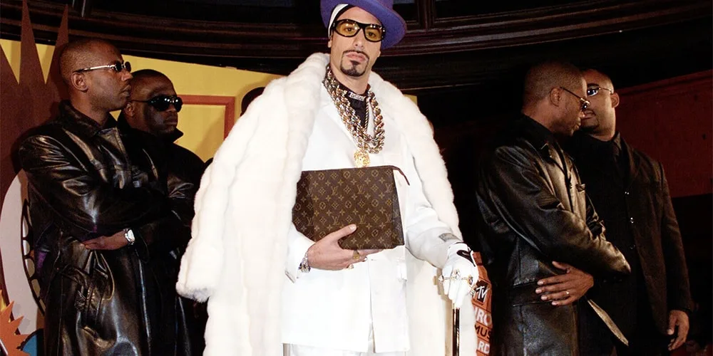 Homme en manteau blanc avec chapeau violet et lunettes, entouré de gardes du corps.
