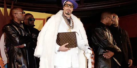 Homme en manteau blanc avec chapeau violet et lunettes, entouré de gardes du corps.