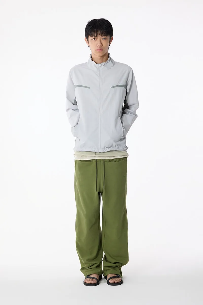 Homme en veste grise et pantalon vert, debout sur fond blanc.