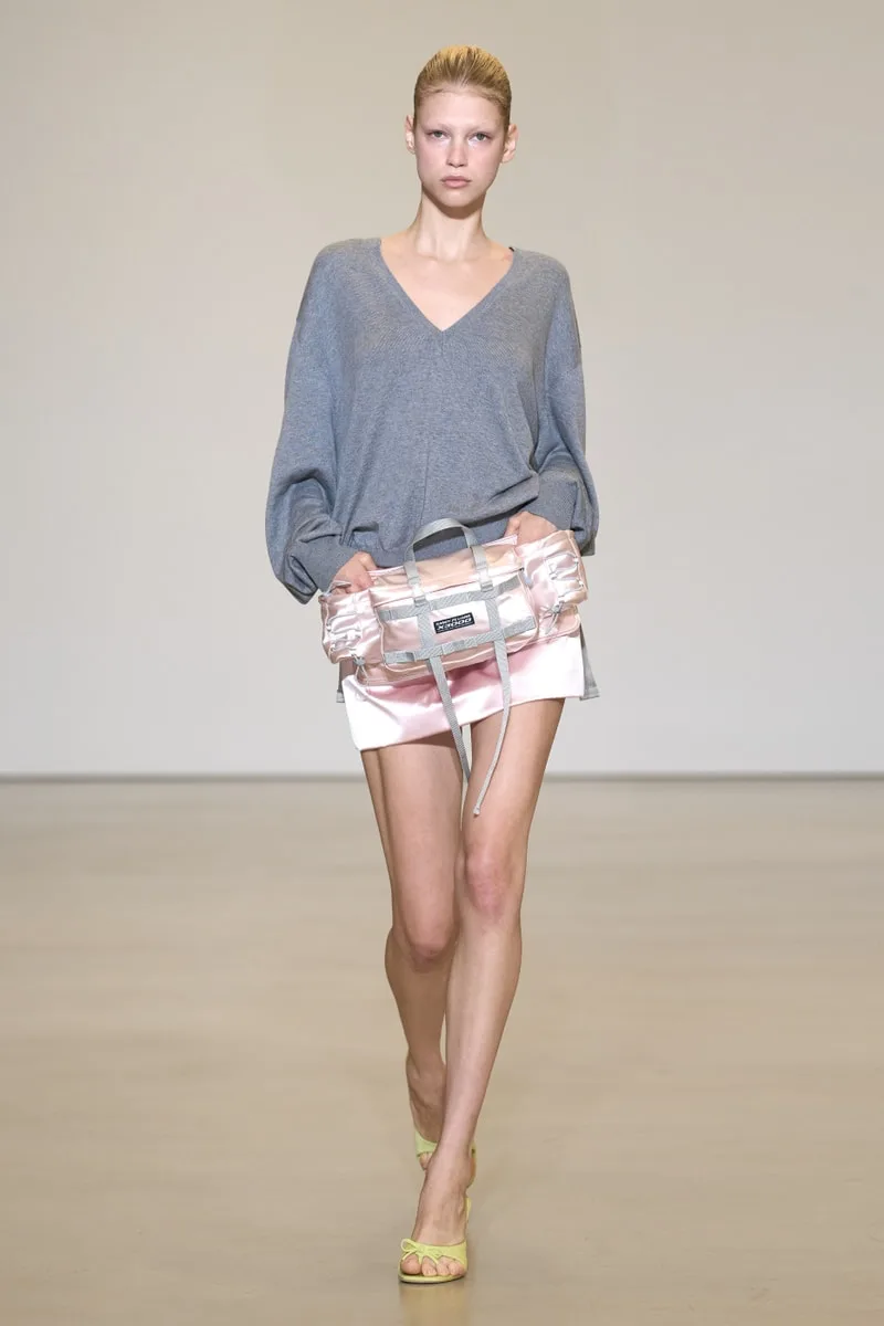 Femme en pull gris et jupe blanche avec sac transparent sur podium.