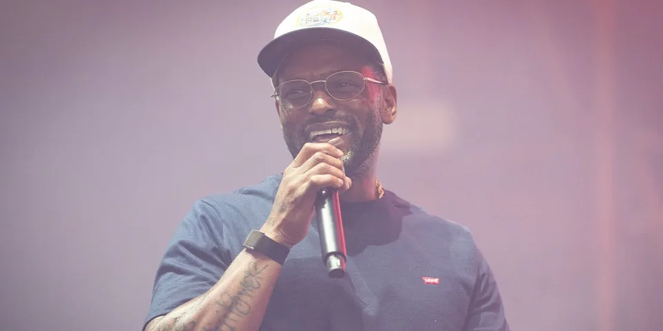 Homme avec une casquette et un micro sur scène, fond rose flou.