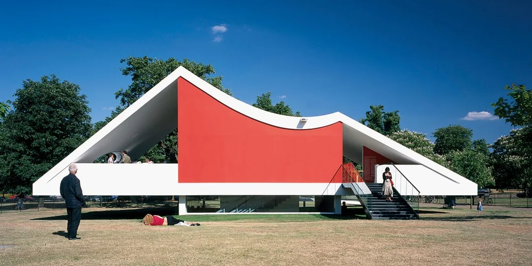 Maison moderne avec toit sculptural et mur rouge, entourée de pelouse et arbres.