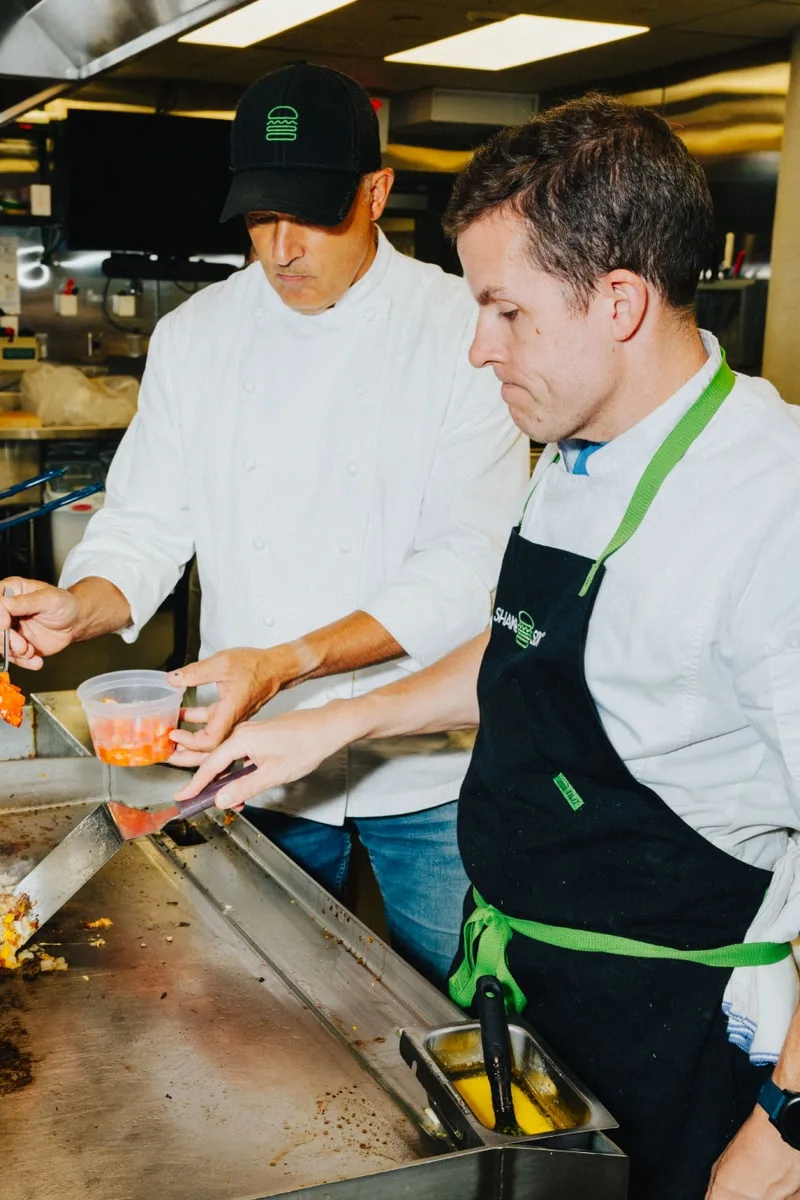 Deux hommes cuisinent dans une cuisine commerciale, portant des tabliers et des chapeaux.
