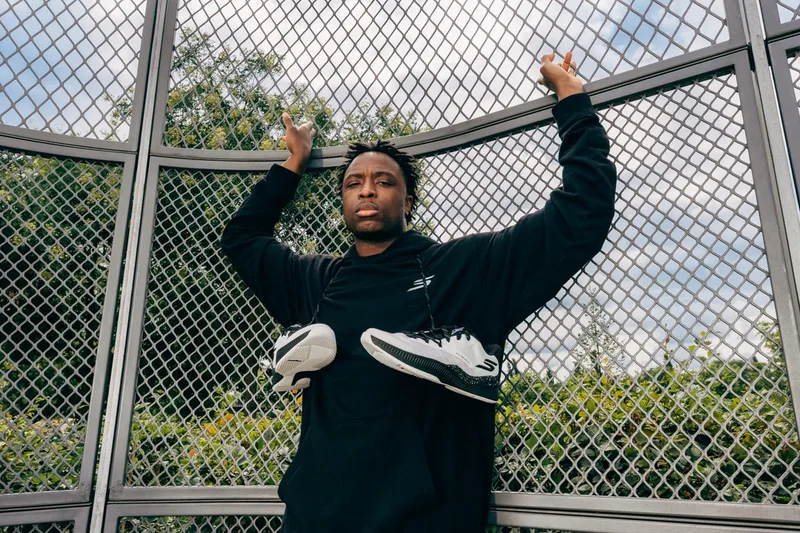 Homme en sweat noir avec des baskets, grillage en arrière-plan.