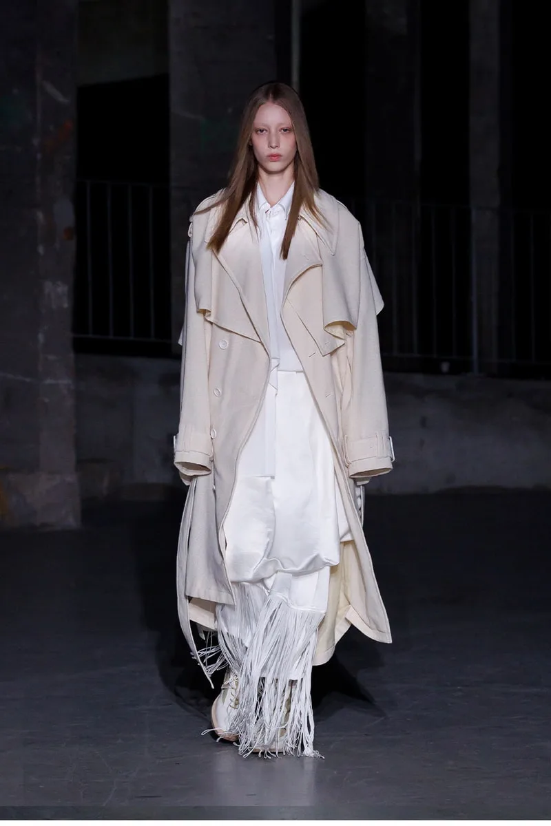 Mannequin en manteau crème et franges blanches sur un podium sombre.