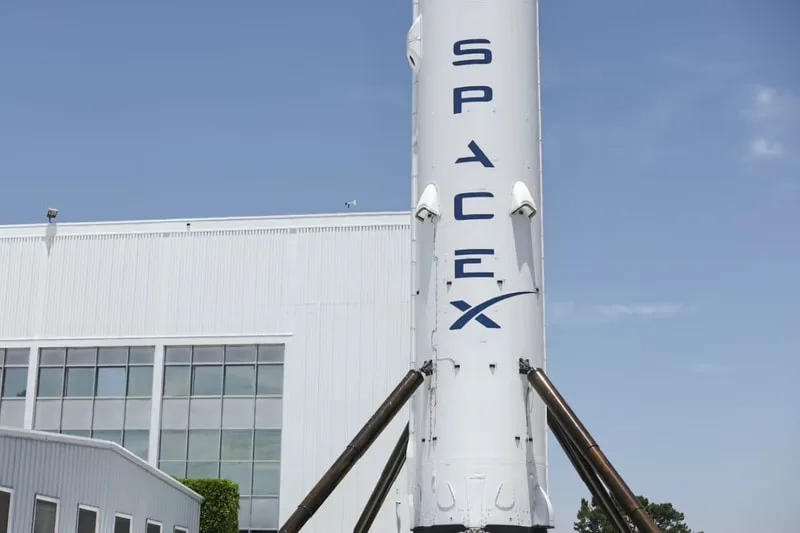 Lanceur SpaceX devant un bâtiment industriel, ciel bleu en arrière-plan.