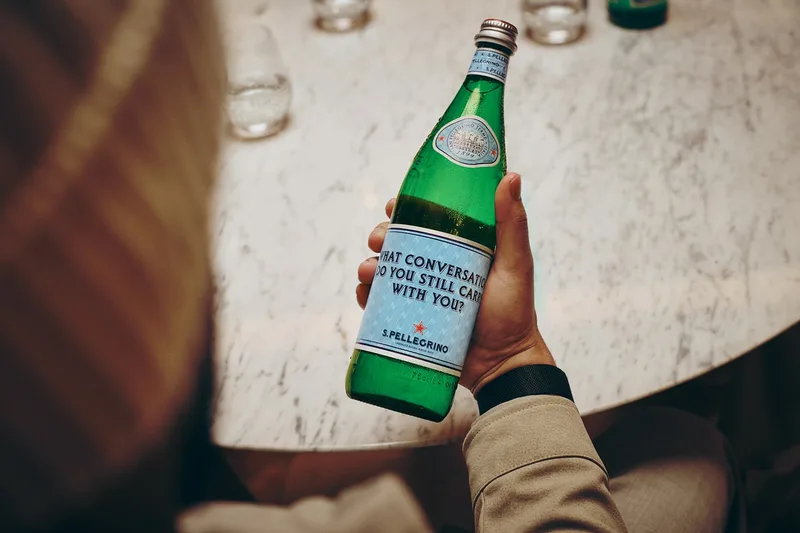 Personne tenant une bouteille d'eau gazeuse San Pellegrino sur une table en marbre.