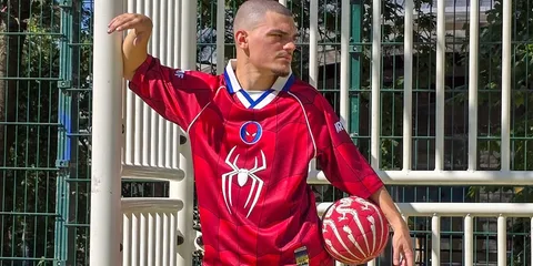 Homme en maillot rouge avec motif araignée et ballon rouge, fond de grille métallique.
