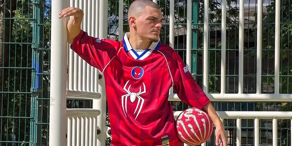 Homme en maillot rouge avec motif araignée et ballon rouge, fond de grille métallique.