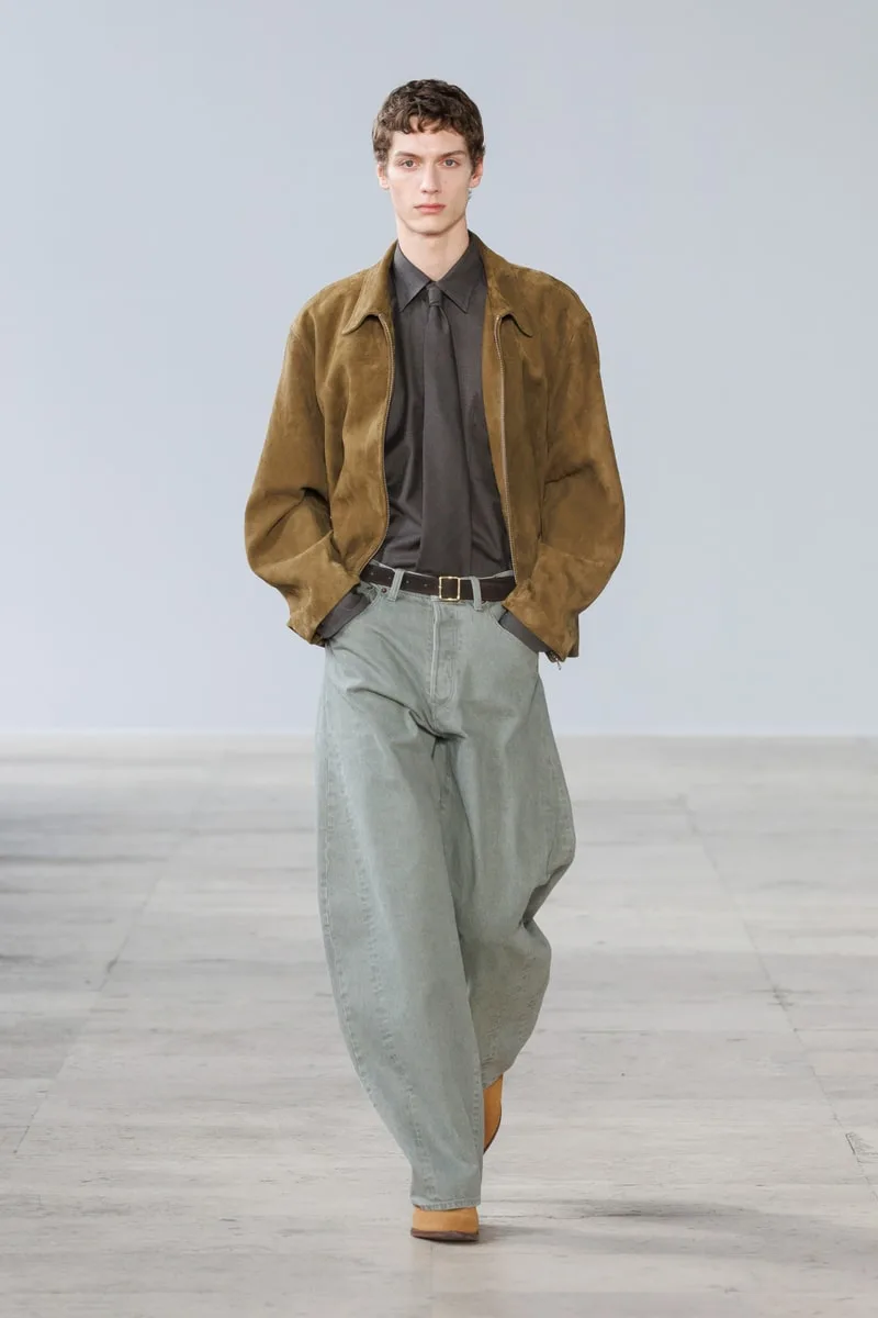 Homme en veste marron et pantalon large gris sur un podium.