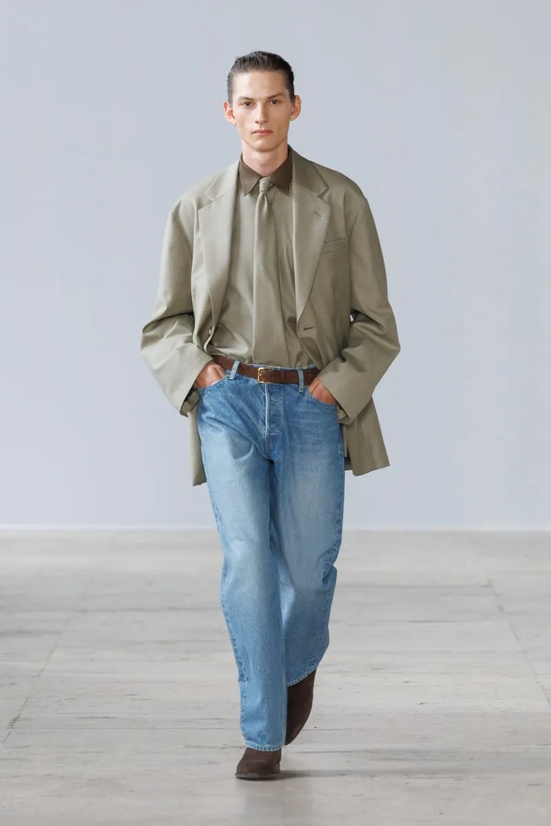 Homme défilant en veste beige et jeans sur un podium.
