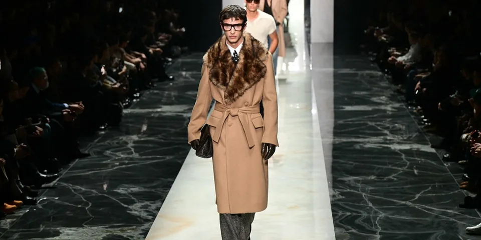 Défilé de mode homme avec manteau long et fourrure, sol marbré.