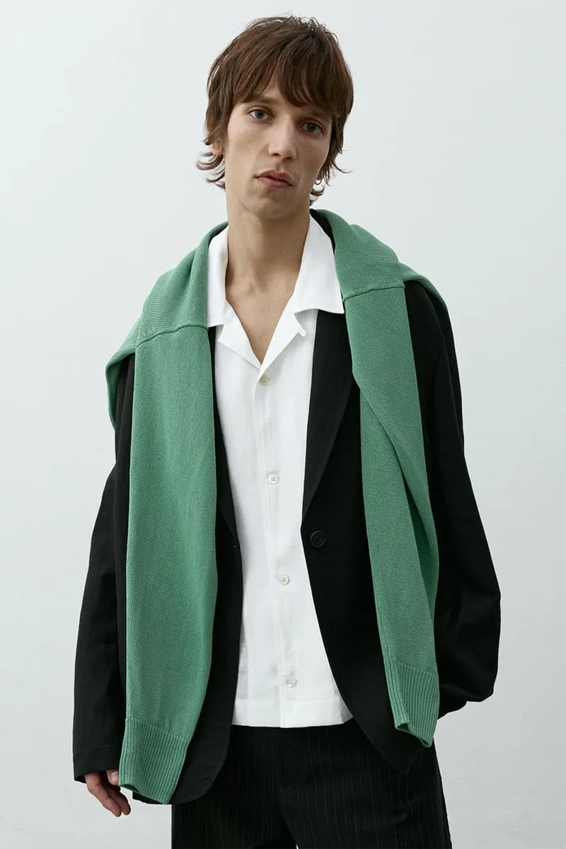 Homme en veste noire et pull vert sur les épaules, chemise blanche dessous.