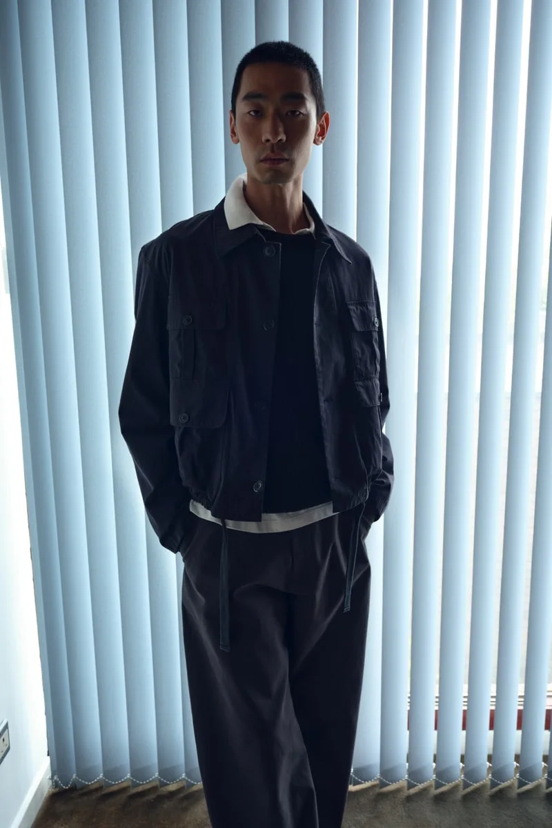 Homme en veste et pantalon noirs devant des stores verticaux.