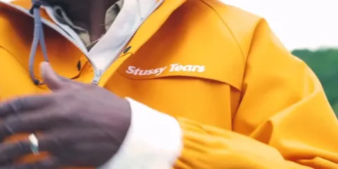 Personne portant une veste orange avec 'Stussy Tears' écrit dessus.