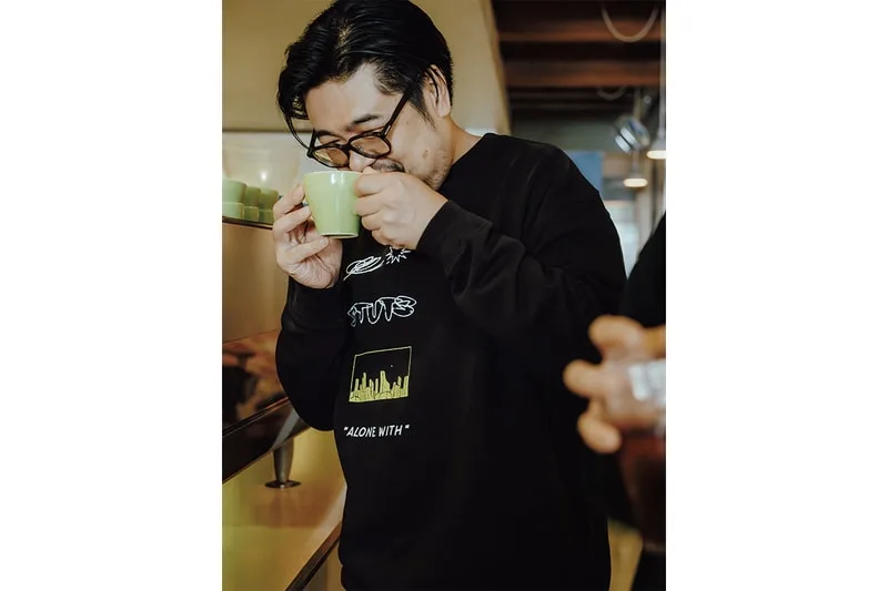 Homme buvant dans une tasse, vêtement noir avec motif graphique vert.