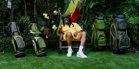 Homme assis avec sacs de golf colorés et végétation tropicale en arrière-plan.