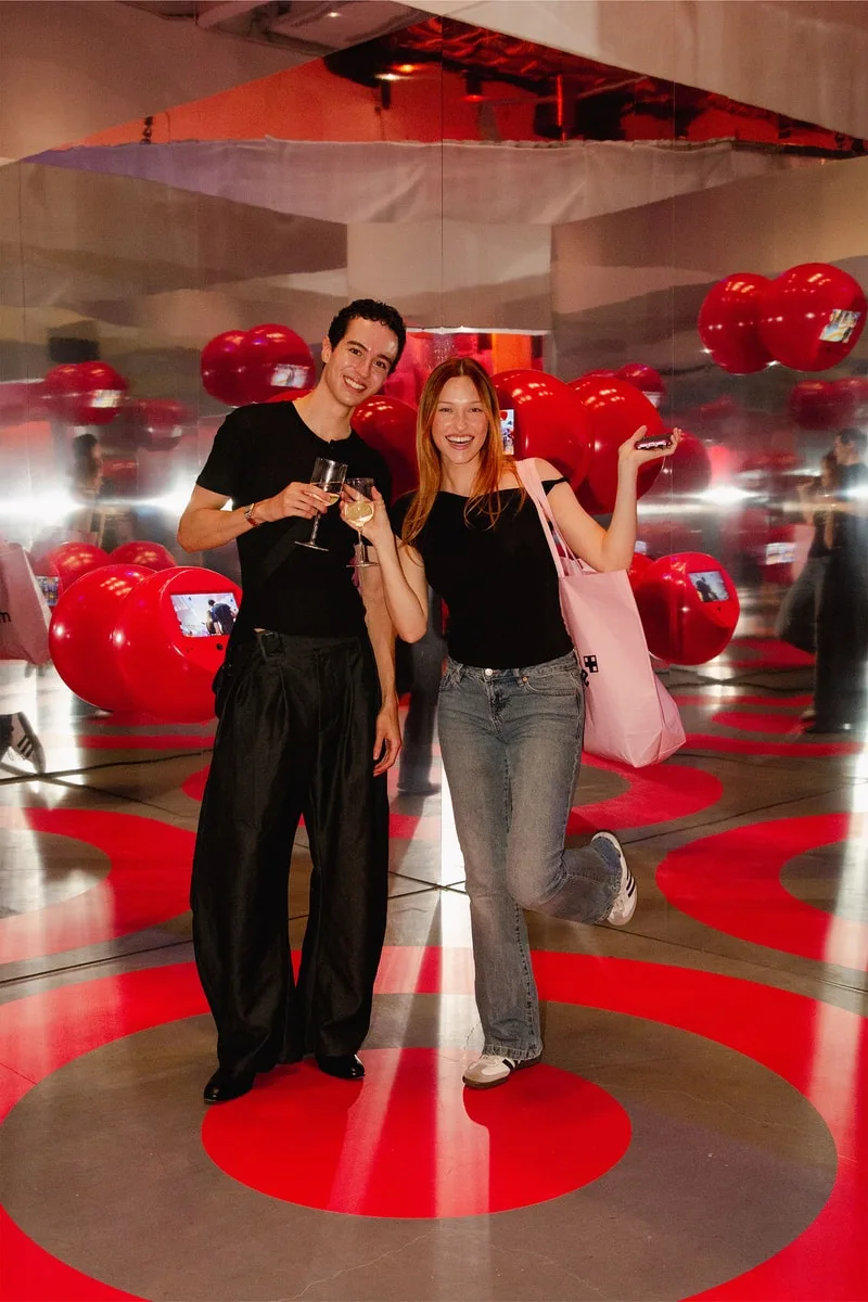 Deux personnes posent dans une salle avec des ballons rouges et un sol à motifs circulaires.