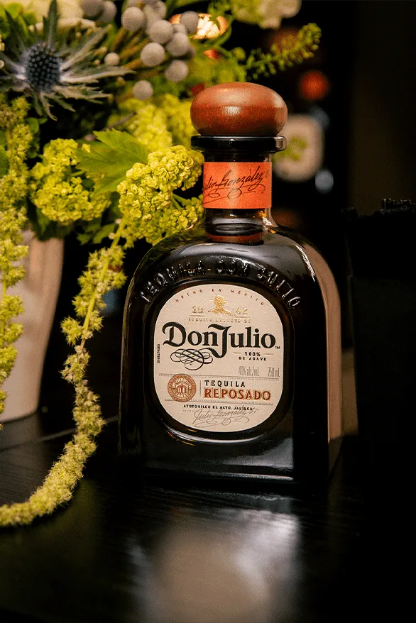 Bouteille de tequila Don Julio Reposado sur une table avec des fleurs en arrière-plan.