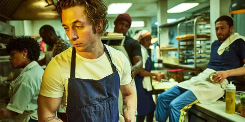 Un chef avec un tablier dans une cuisine occupée avec plusieurs personnes.