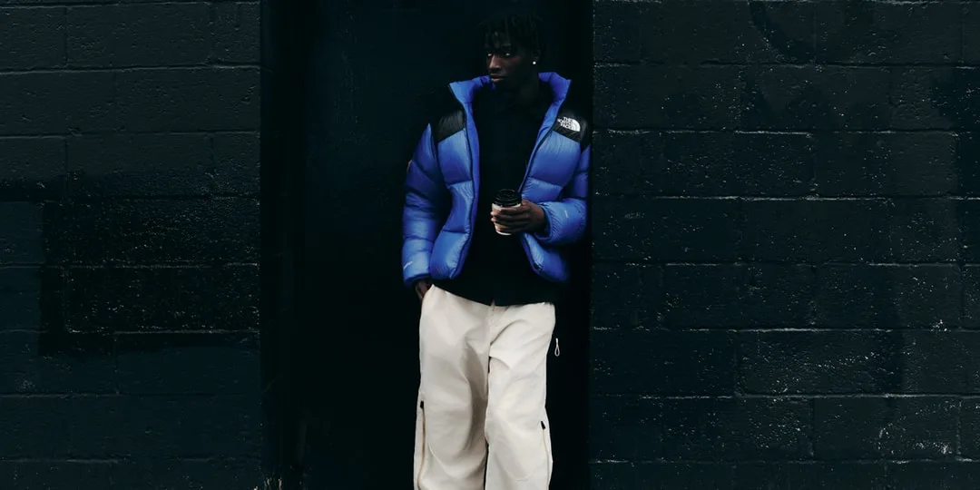 Un homme en doudoune bleue et pantalon blanc, adossé à un mur sombre.