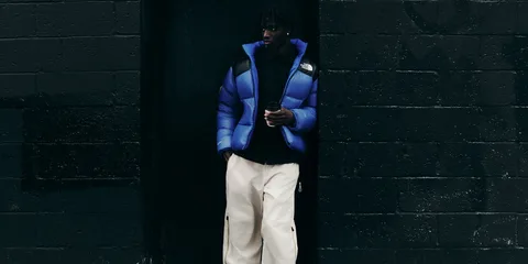 Un homme en doudoune bleue et pantalon blanc, adossé à un mur sombre.