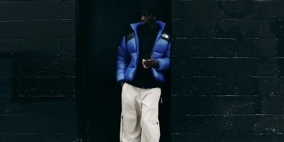 Un homme en doudoune bleue et pantalon blanc, adossé à un mur sombre.