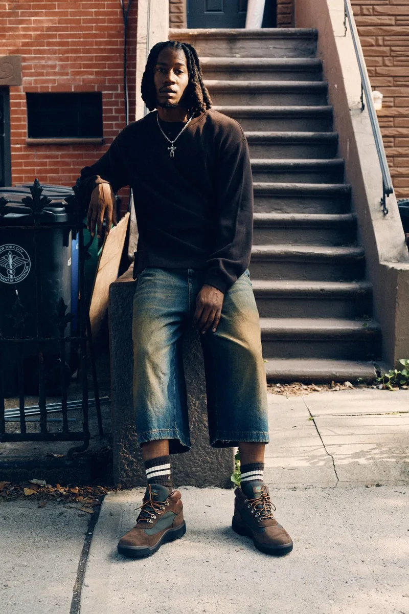 Homme assis devant des marches, portant un pull et un jean large.
