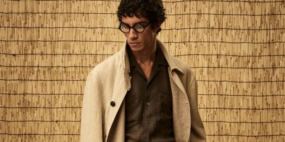 Homme avec lunettes et manteau beige devant un fond en bambou.