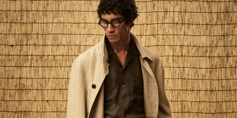 Homme avec lunettes et manteau beige devant un fond en bambou.