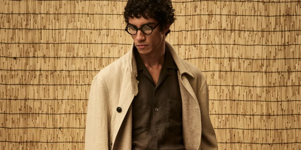 Homme avec lunettes et manteau beige devant un fond en bambou.