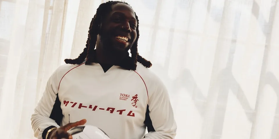 Homme souriant en maillot blanc, tenant un ballon, lumière naturelle en arrière-plan.