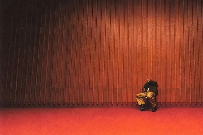 Personne assise contre un mur en bois dans une pièce rouge, isolée et méditative.