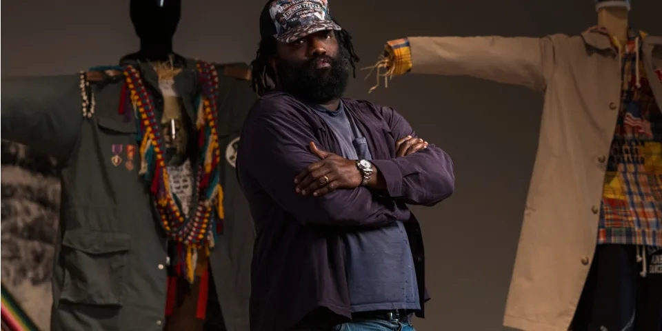 Homme avec barbe, vêtements colorés, debout devant mannequins.